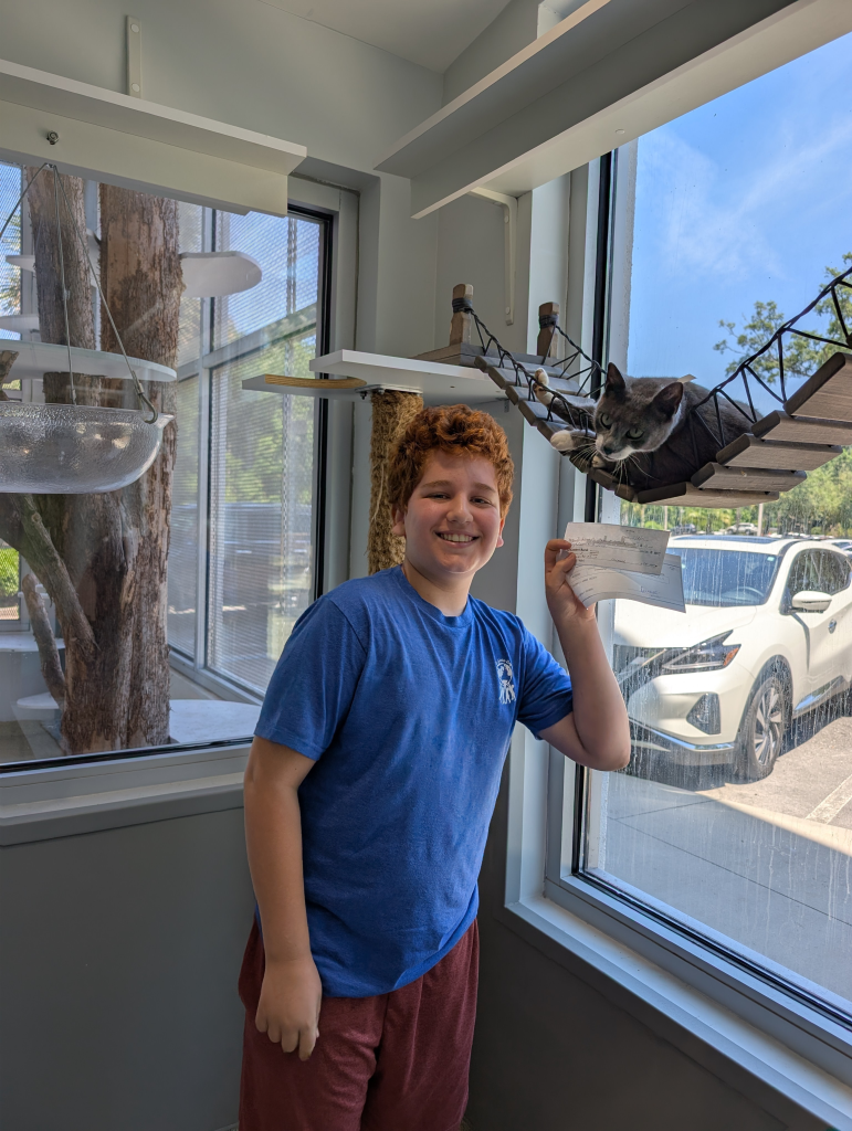 Koren standing next to Eve the cat, who is lounging on a wooden bridge hanging in one of JHS's group cat rooms. Koren is holding checks (donations to JHS).