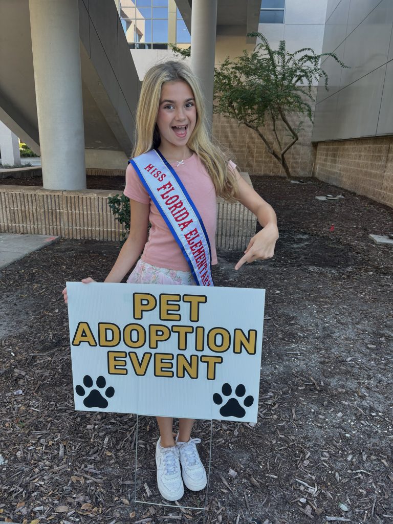Addie posing behind signage with her Miss Florida Elementary sash at the Pet Adoption Festival hosted at the Prime Osborn Convention Center in Downtown Jacksonville.
