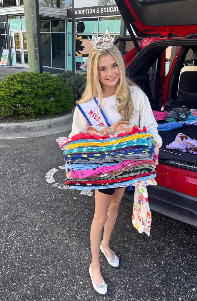 Addie is pictured next to a red SUV holding a stack of fleece blankets to be donated to JHS outside by the Adoption and Education Center main entrance.