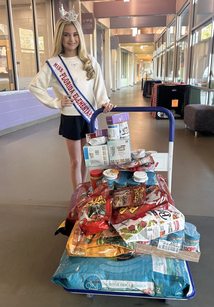 Addie posing behind one of the carts full of donations with her Miss Florida Elementary 2026 crown and sash. The donations include wet dog food, wet cat food, kitten formula, treats, peanut butter, and kitten food.