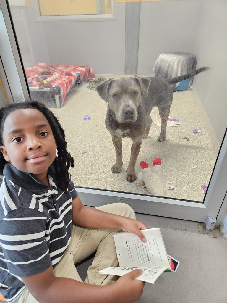 Nehemiah is pictured sitting down crisscrossed reading aloud to Hopper, a 4 year old dog standing at the door of his suite kennel.